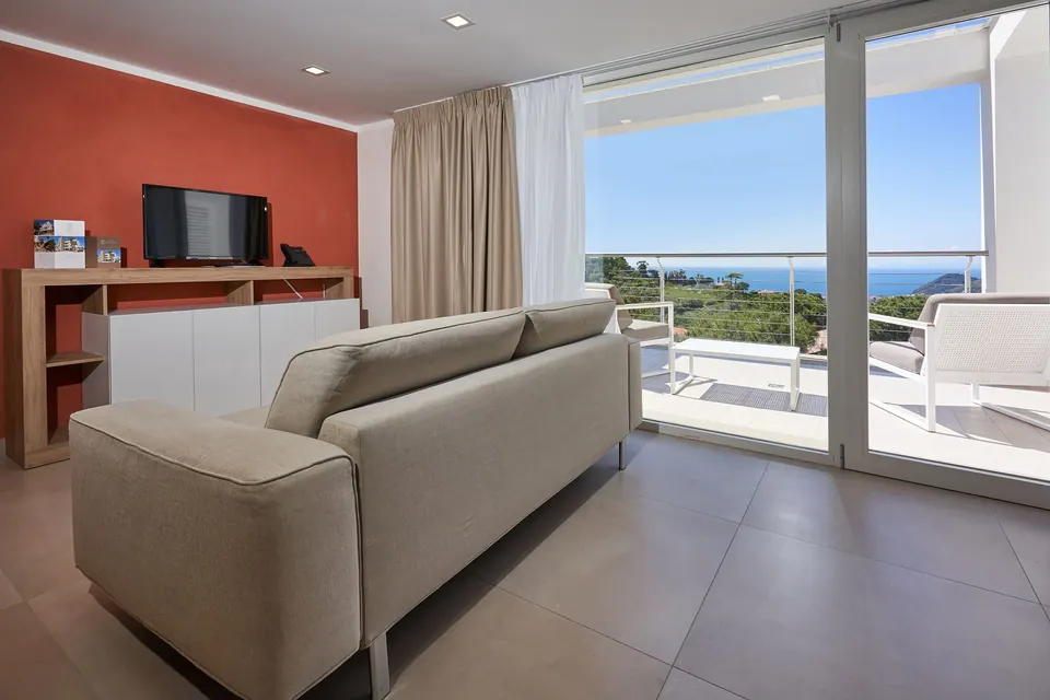 Living area of the quadruple room with sofa, round table and glass doors facing the sea
